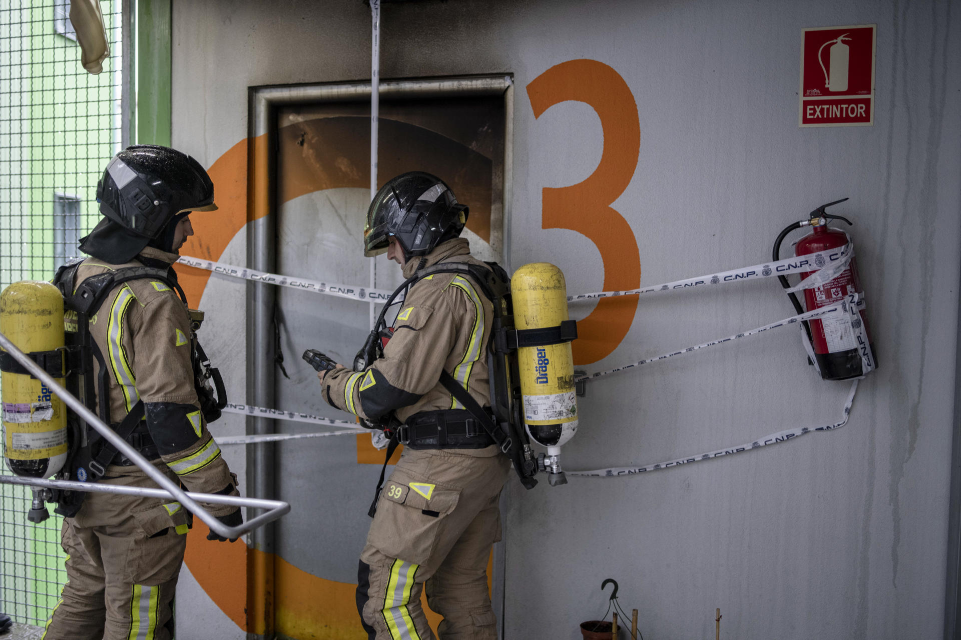 Dos bomberos realizan una medición en la puerta de la vivienda incendiada este sábado en Ourense en la que han muerto dos mujeres de 50 y 71 años. EFE/ Brais Lorenzo