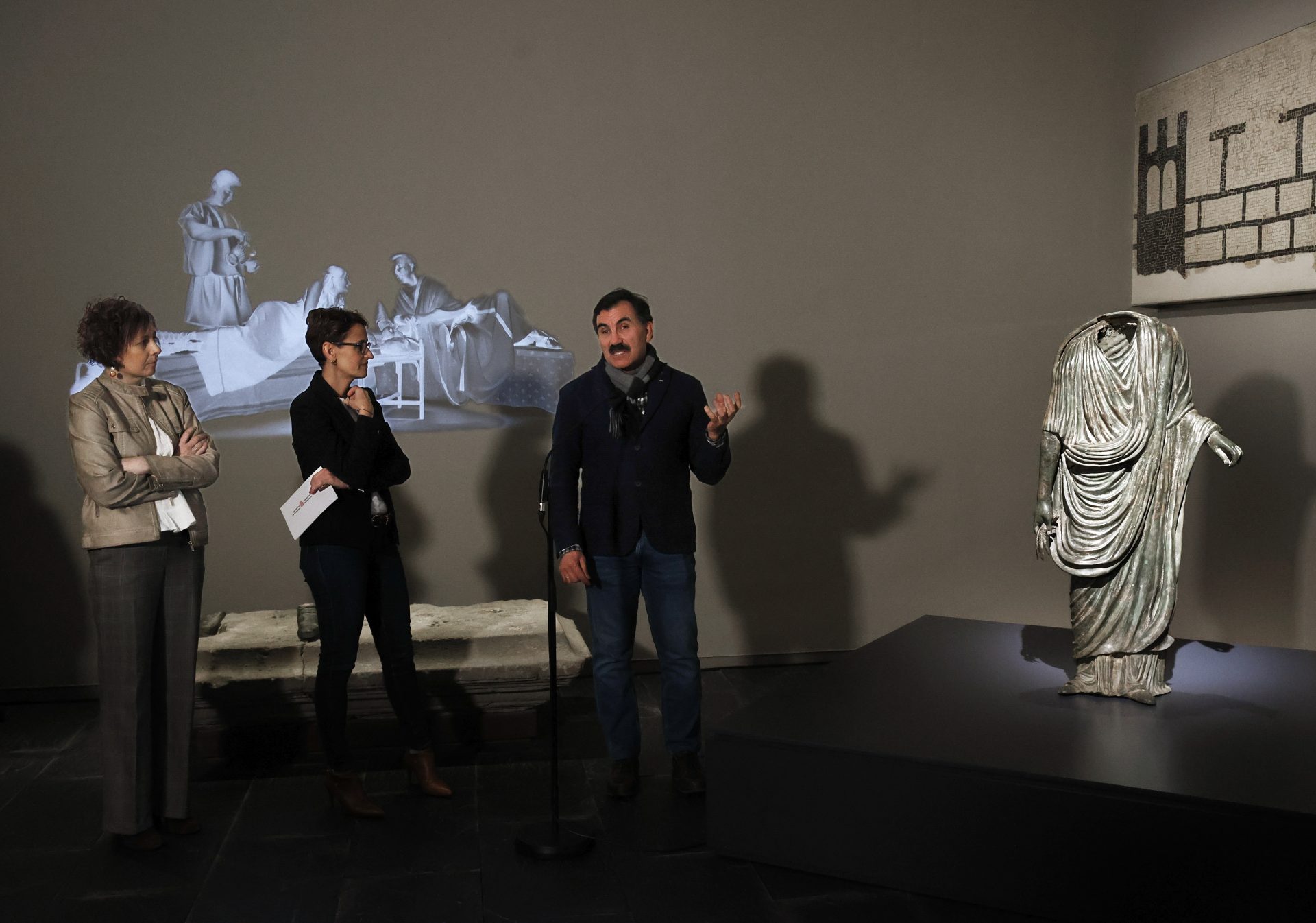 El arqueólogo Jesús Sesma explica a la presidenta de Navarra y la consejera de Cultura detalles de la escultura “El togado de Pompelo”