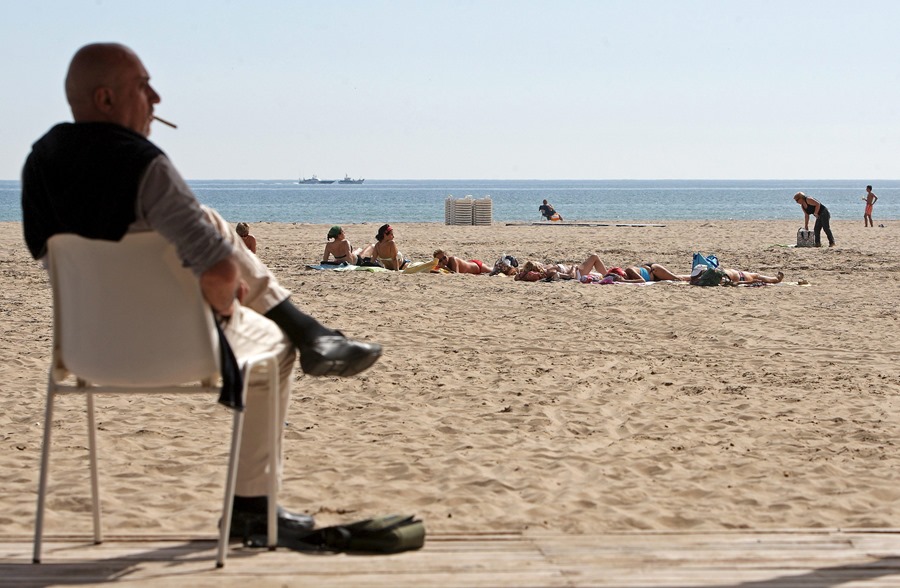 Un grupo de mujeres toma el sol este mediodía en la playa de Las Arenas de Valencia mientras un hombre fuma un cigarrillo sentado en le paseo marítimo.
