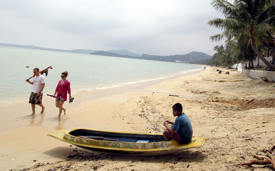 Un tailandés descansa en su barca mientras dos turistas pasean por una playa en la isla de Koh Samui. 