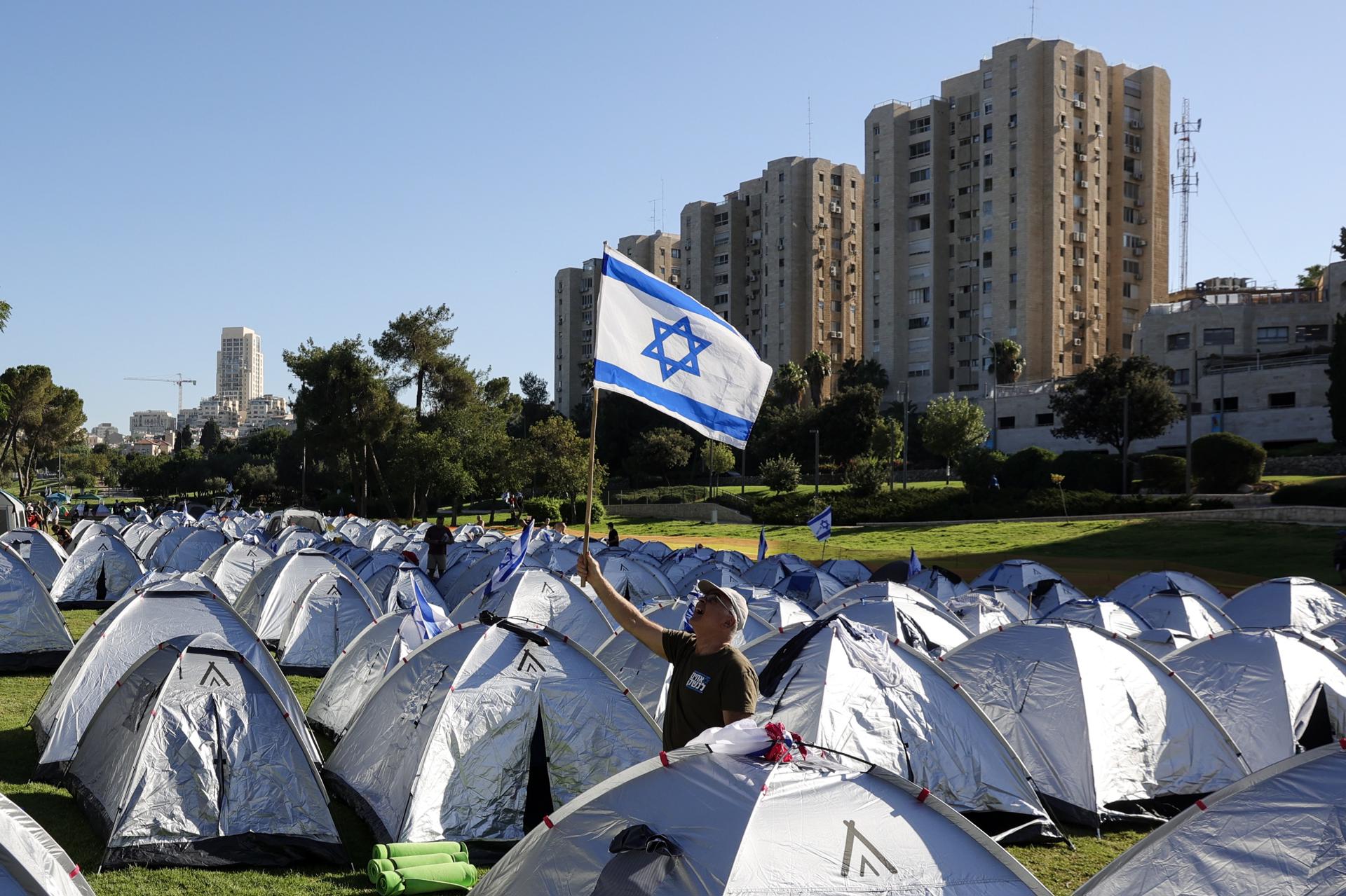An anti-government protester raises a Israeli flag among tents after spending the night in a tent camp at Sacher Park, near the Israeli Knesset, following a four-day protest march to Jerusalem against the government's planned justice system reform, in Jerusalem, 23 July 2023. EFE/EPA/ABIR SULTAN

