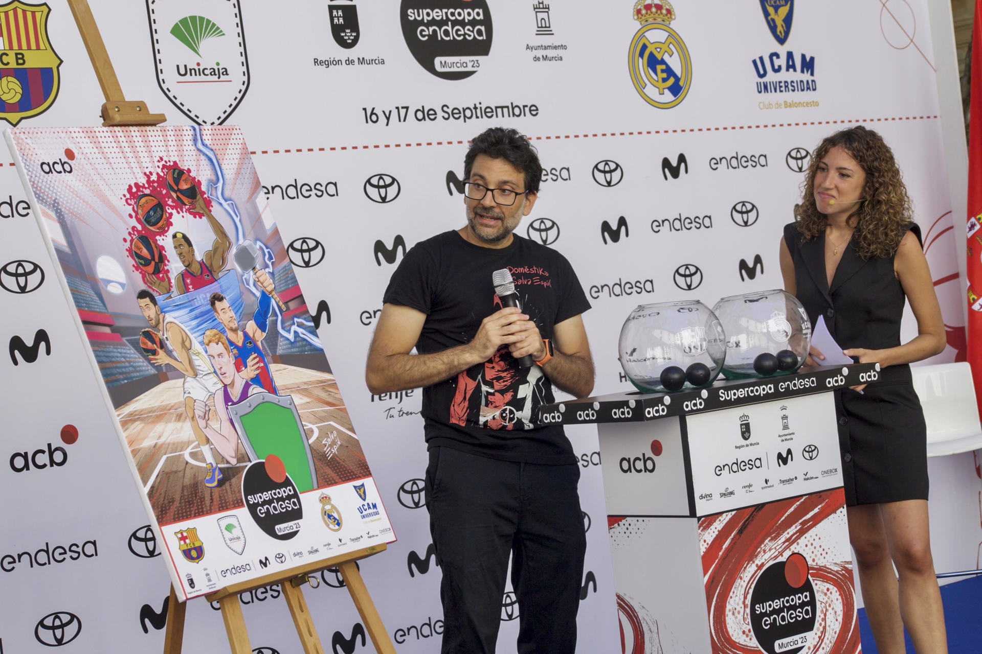 El dibujante murciano de Marvel y creador del cartel de la Supercopa, Salva Espín (i), muestra su obra este martes en el Palacio de San Esteban de Murcia durante el sorteo de emparejamientos de la Supercopa. EFE/ Juan Carlos Caval
