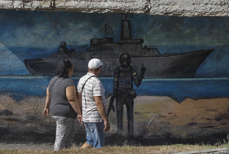 Two people walk past a mural depicting a Ukrainian soldier pointing at a Russian warship, in kyiv, Ukraine