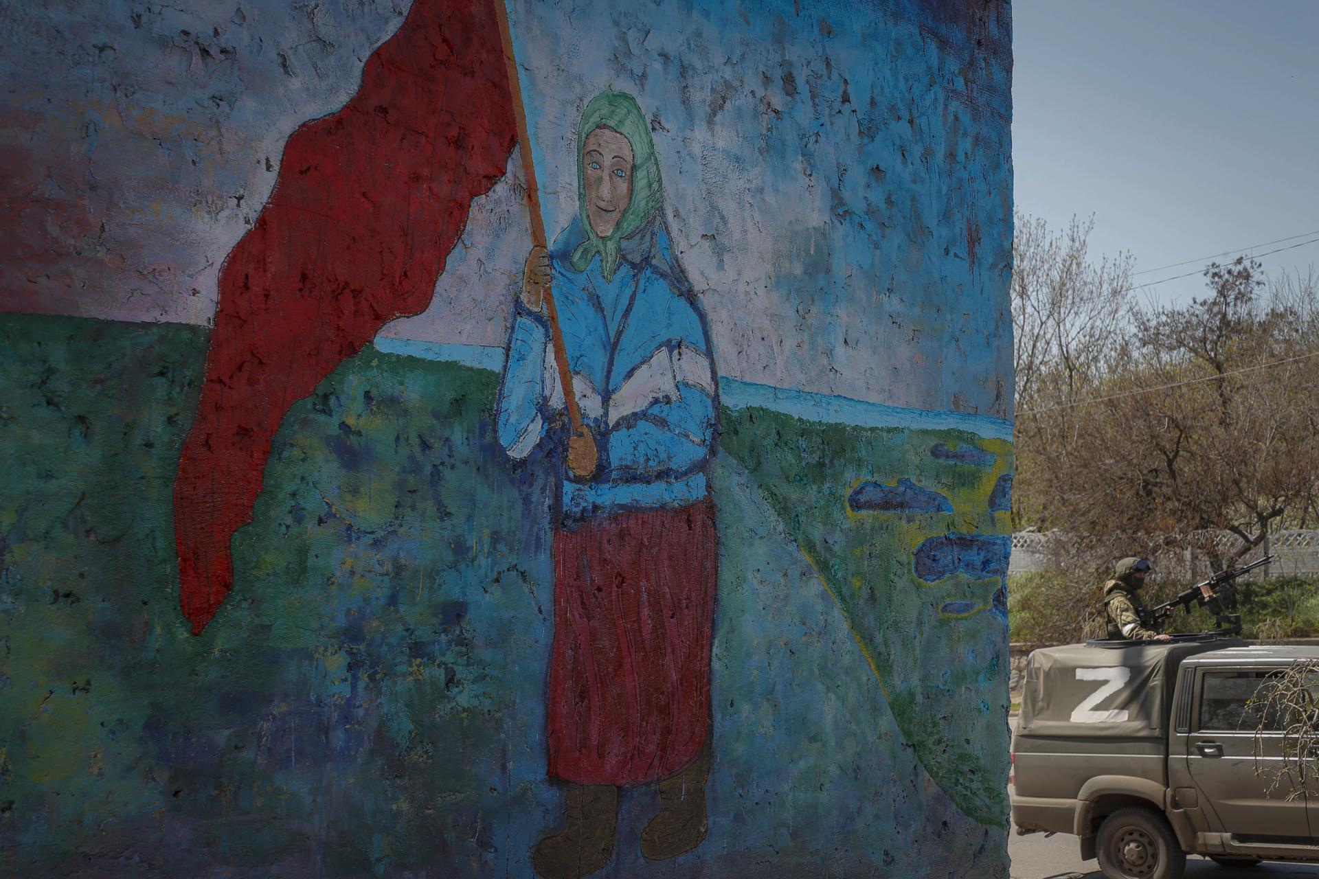 A picture taken during a visit to Mariupol organized by the Russian military shows Russian serviceman guards behind the painting of Ukrainian old woman with replica of the USSR red flag in downtown of Berdyansk, Ukraine, 30 April 2022 (Issued on 01 May 2022). EFE-EPA FILE/SERGEI ILNITSKY
