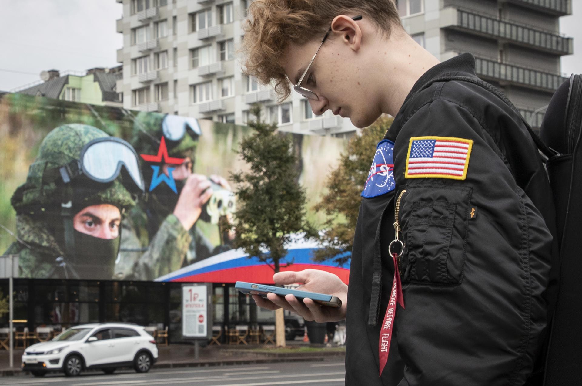 A young man wearing a shoulder patch depicting a US flag waits for a bus near a billboard advertising military conscription in Moscow, Russia, 18 September 2023. EFE-EPA/YURI KOCHETKOV
